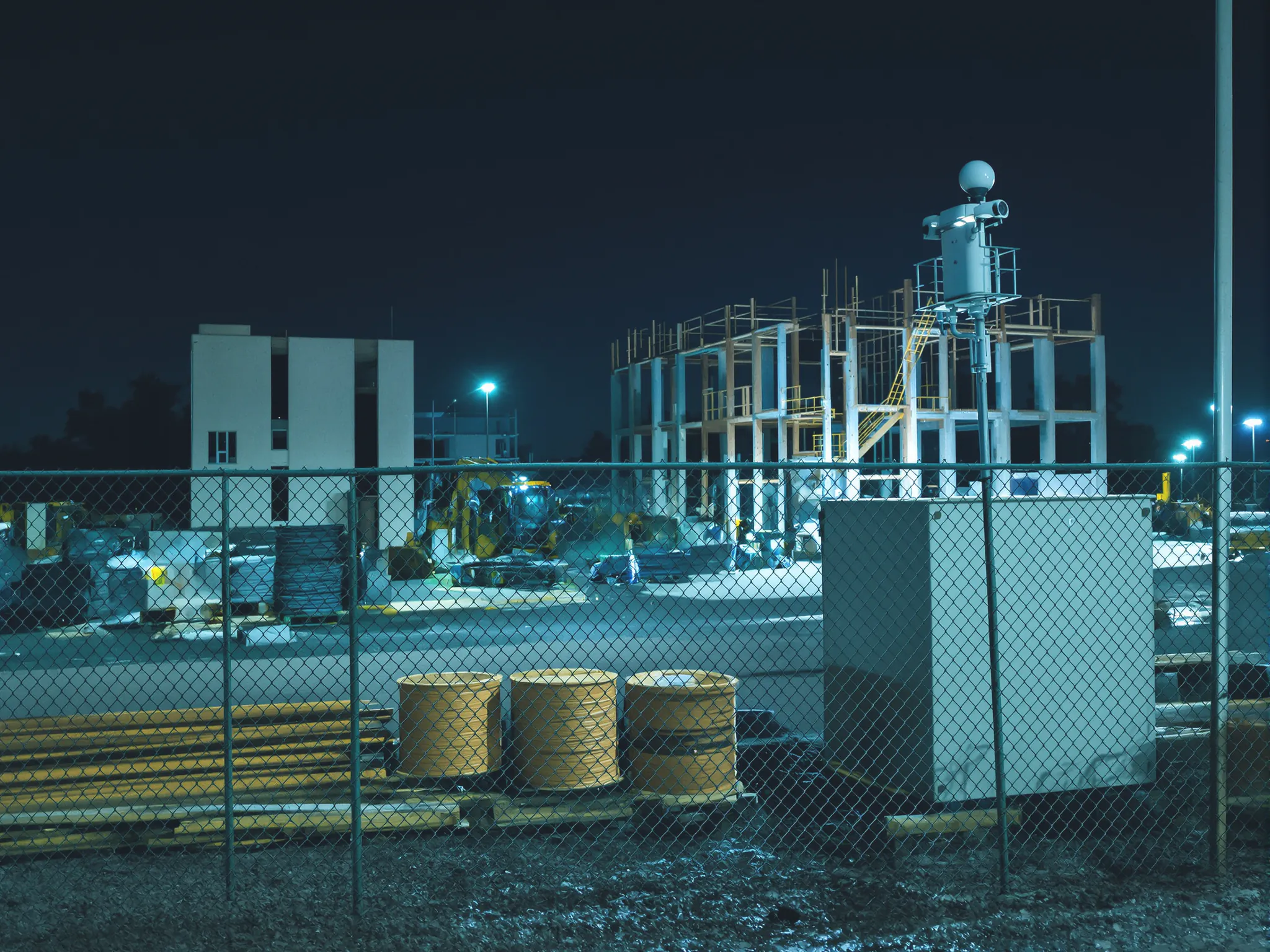 A fenced construction site at night: cable reels and stacked lumber in the materials staging area, a steel storage box and a camera pole, with a half-built frame behind.